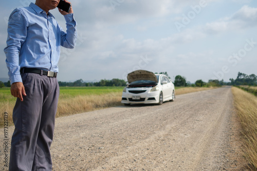 A man is using his cell phone to call for help because his car broke down on a dirt road far from home. He ran out of fuel during the journey in this energy-scarce situation.