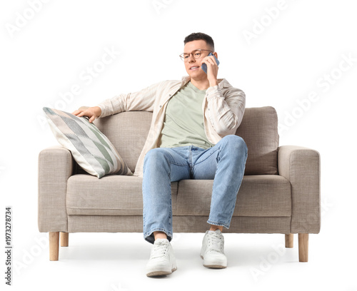 Fotografie Young man talking by mobile phone on grey sofa against white background