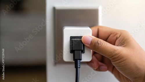 Hand plugging a black charger cable into a white electrical outlet on a beige wall.