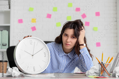 Tired young businesswoman with clock at her workspace in office. Deadline con...