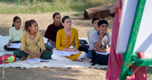 Indian adult female lady talk speak teach to villager group day under tree girl boy sitting on mat do study look board learn at open class field rural area place