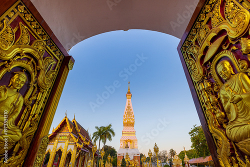 Foto Golden Pagoda of Wat Phra That Phanom through Ornate Temple Doorway at Twilight, Nakhon Phanom, Thailand,A stunning wide-angle view of the iconic Phra That Phanom pagoda, a sacred Buddhist landmark