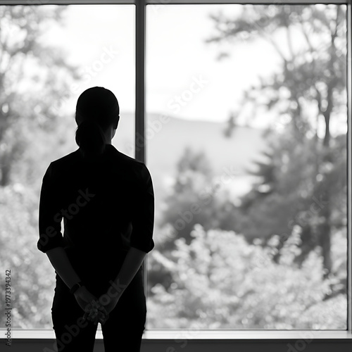 Person silhouette standing looking out of a large window with hands clasped behind the back in black and white