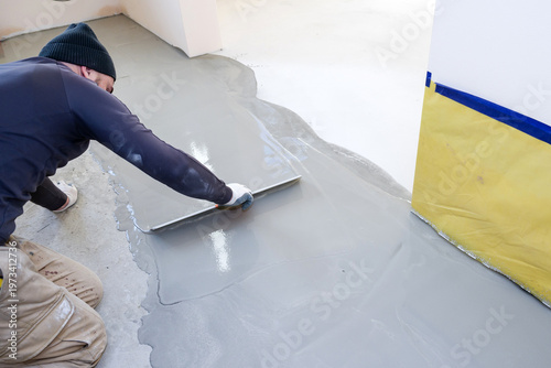 Worker applies fresh cement to the self-leveling floor in a construction area during the day with tools and protective gear used for flooring tasks