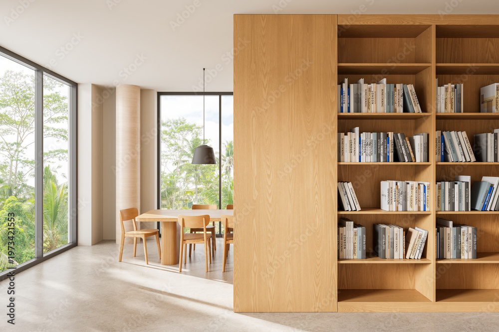 Naklejka premium Bright minimal interior with wooden dining table, chairs, and large bookshelf beside expansive windows overlooking lush greenery and soft daylight..