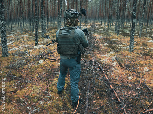 Rear view of special forces operator with night vision, tactical vest and rifle standing in a wild forest during twilight scout mission.