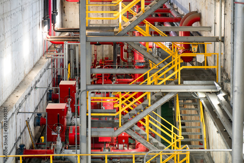 An intricate view of a multi-level industrial facility featuring bright red piping, high-pressure valves, and yellow metal safety stairs. The image highlights a sophisticated mechanical engineering sy