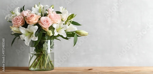 A bouquet of delicate pink roses and white lilies arranged in a clear glass vase on a wooden surface against a light gray background.
