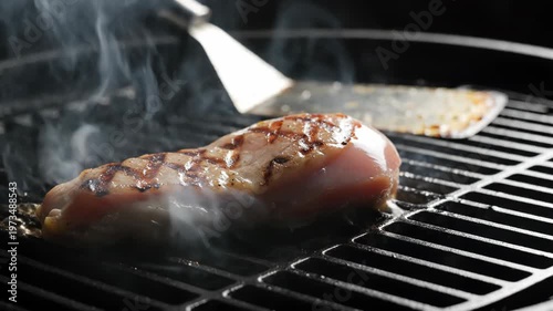 Close-up of a raw chicken breast cooking on a hot black metal grill with visible charred grill marks and rising white smoke, a metal spatula sits in the background of the scene.