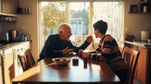 Candid documentary-style photo of elderly grandfather and teenage grandson laughing together at a kitchen table