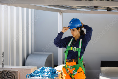 Female industrial technician putting on blue hard hat and safety harness for rooftop work, Professional maintenance worker preparing safety equipment for air conditioning service