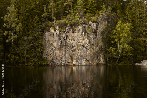 Obraz na płótnie A striking granite cliff face rises dramatically from the edge of a calm, dark boreal forest lake in Norway, with dense pine and birch trees and a clear, detailed reflection on the still water