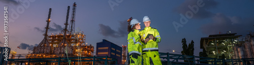 Two petroleum engineers discussing project and pointing direction with clipboard and radio communication at oil refinery site during sunset teamwork planning and industrial energy infrastructure.