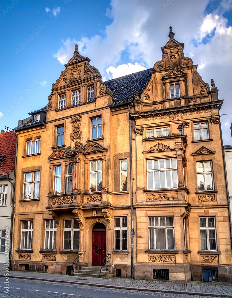 Fototapeta premium An ornate, multi-story building with symmetrical design, featuring detailed stonework, windows, and a red door, under a blue sky