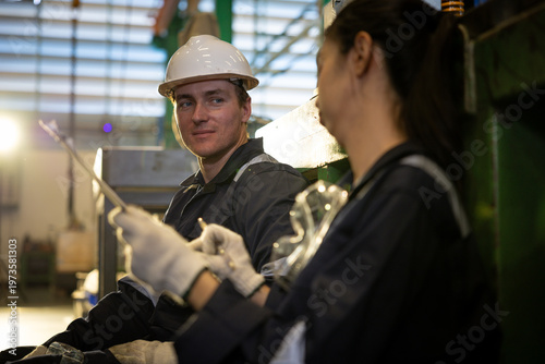 Confident smiling male factory worker wearing dark uniform and white safety helmet collaborating with female engineer during industrial teamwork inspection process
