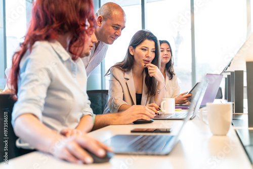 A diverse group of professionals gathered around a laptop, engaged in a meeting or discussion, with a focus on teamwork and collaboration in a modern office environment.