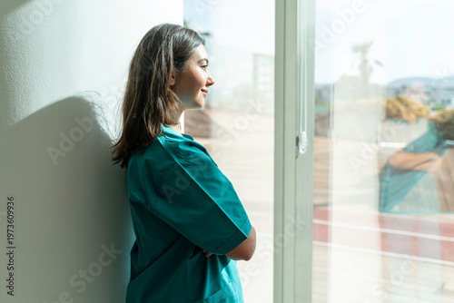 A female nurse or doctor in green scrubs stands by a hospital window, looking outside