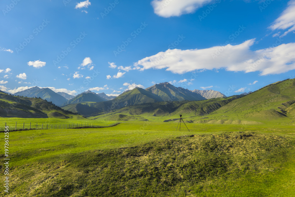 Fototapeta premium Tripod with camera equipment set against a backdrop of green rolling hills and clear blue sky. Peaceful scene captures the essence of nature's beauty in Kyrgyzstan.