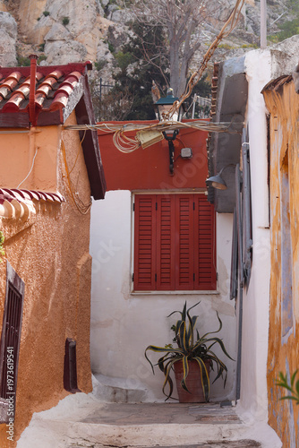 Charming narrow alley in Anafiotika, Athens, with warm colored walls, wooden shutters and potted plant, capturing authentic Mediterranean old town atmosphere.