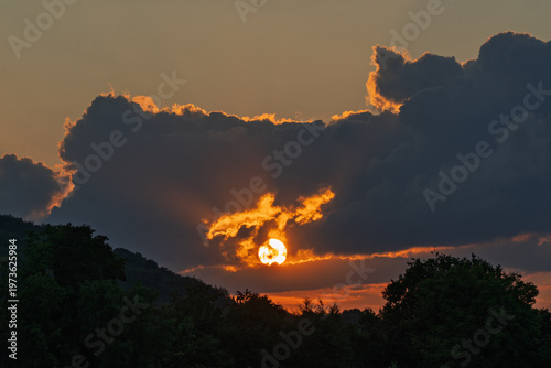 Sunset through clouds over a wooded hilly landscape.