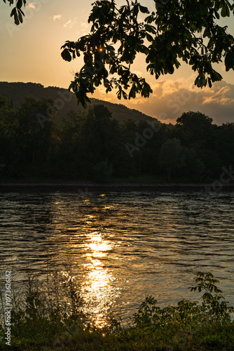 Shortly after sunset over a wooded hilly landscape behind a riverbank.