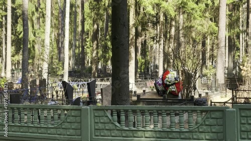 People visit a cemetery in a forest setting to lay flowers and pay respect. The sun shines through the trees as visitors approach the memorials quietly.
