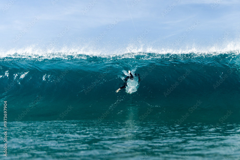 Obraz premium A lone surfer duck dives beneath a towering turquoise wave, cutting through white spray as the ocean swells overhead under a clear, calm sky.