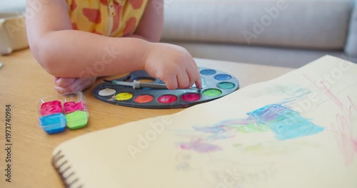 Creative toddler dips a finger into a colorful watercolor paint palette at a wooden table indoors, focusing on early childhood development, creative sensory play, and hands-on learning.