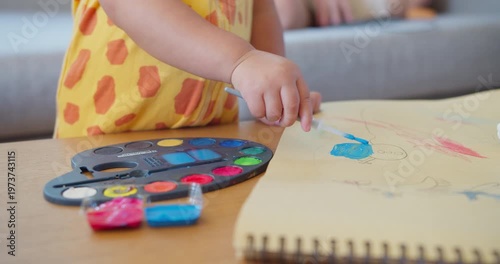 Unrecognizable toddler carefully paints colorful artwork on a sketchbook at a living room table, focusing on early childhood development, creative expression, and fine motor skills.
