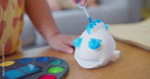 Child carefully paints a plaster fish figurine with a blue brush on a wooden table, focusing on early childhood development, creative play, and artistic expression.