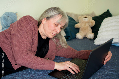 Mature woman working on laptop lying on bed at home office