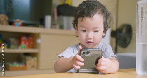 Asian toddler attentively holds and watches a smartphone at a wooden living room table indoors, focusing on early digital literacy, childhood screen time management, and modern technology.