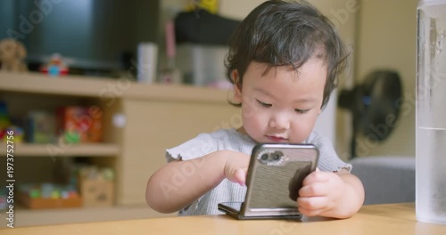 Asian toddler watches digital content on a smartphone in a bright living room, focusing on early childhood technology, modern screen time, and interactive home entertainment.