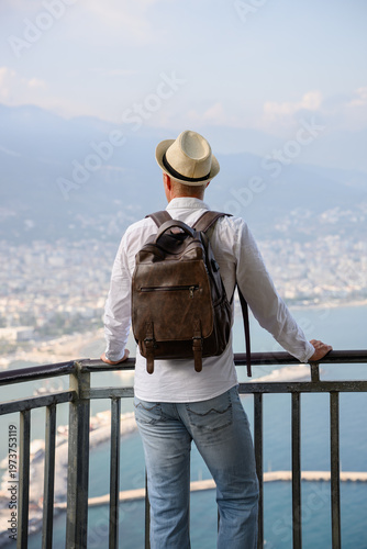 Male traveler in straw hat with backpack looking at scenic coastal city view from observation deck. Summer vacation travel discovery, solo adventure lifestyle, mediterranean tourism, freedom, holidays