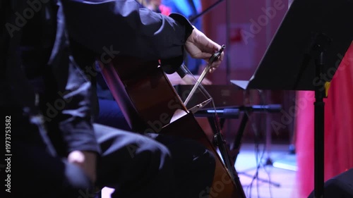 Musician in black formal attire draws horsehair bow across strings of wooden cello while seated on stage near music stand during orchestral performance