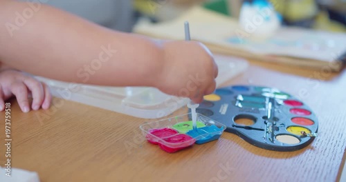 Young toddler dips a paintbrush into bright blue paint at a wooden table during an art activity, focusing on early childhood education, fine motor skills, and creative expression.