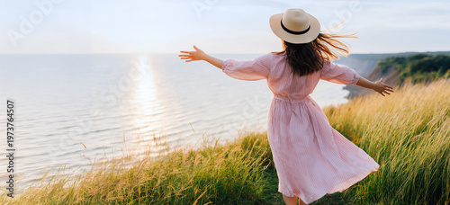 Young woman in straw hat and pink dress standing on grassy cliff with arms outstretched facing sea at sunset with golden light and horizon view, freedom and serenity concept