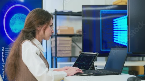 professional woman with glasses in modern AI development office focuses intently on coding at laptop while viewing large blue AI Assistant screen with glowing neural brain graphic, symbolizing machine