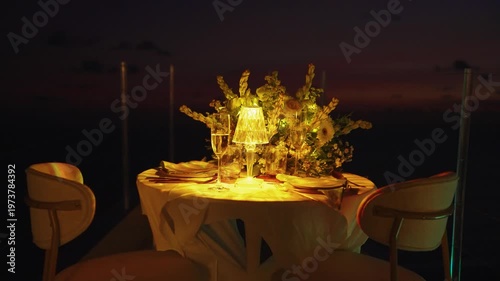 Table for romantic dinner at night served on the outdoor terrace of restaurant with sea sunset view, illuminated by a warm glowing lamp. Elegant decorated setting for dating