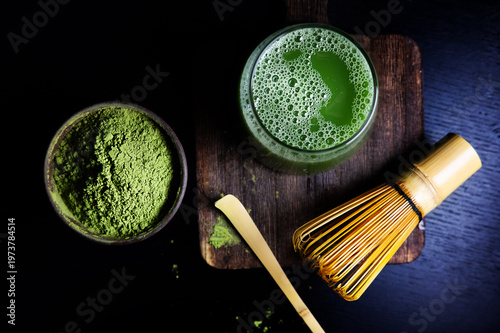 Cup of matcha green tea with foam alongside matcha powder in a bowl, bamboo spoon and whisk.