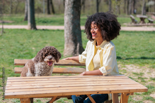 Woman sits at a table in a park with her dog enjoying a sunny day outdoors during spring time with trees in the background