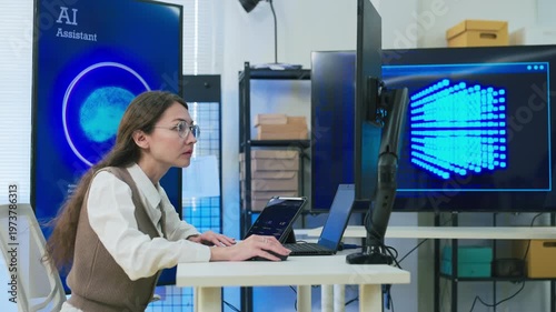 professional woman with glasses in modern AI development office focuses intently on coding at laptop while viewing large blue AI Assistant screen with glowing neural brain graphic, symbolizing machine