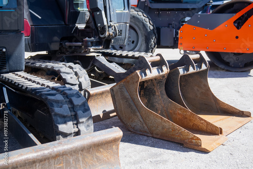 Foto Mini excavator tracks with two bucket attachments on worksite surface, reflectin