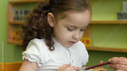 Little girl learning letters with teacher using wooden blocks at table. Teacher hands pointing with pencil, child repeating sounds and looking attentive. Early education, preschool learning concept.