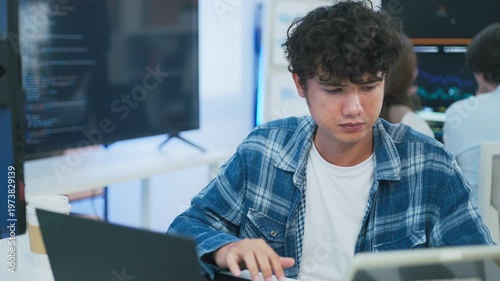 focused young male developer with curly hair in blue plaid shirt intensely codes on laptop in modern AI office, surrounded by multiple monitors displaying code neural networks and data graphs	