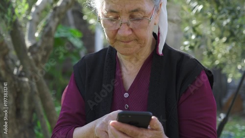 Elderly woman with glasses and headscarf using her smartphone in a garden, smiling and looking happy while scrolling and typing on the touch screen outdoors