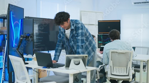 focused young male developer with curly hair in blue plaid shirt intensely codes on laptop in modern AI office, surrounded by multiple monitors displaying code neural networks and data graphs	