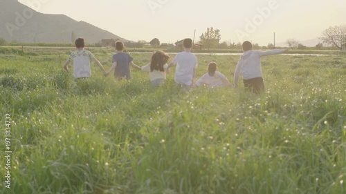 Group of children holding hands running together through a green grass field at sunset. Back view of friends having fun in nature, enjoying a happy childhood outdoors