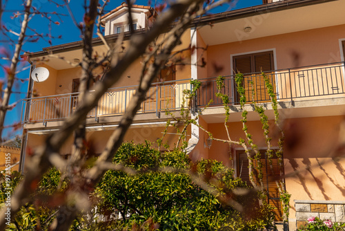 Mediterranean house in Medulin with balcony and shutters, framed by tree branches in soft focus, captured in warm sunlight, showcasing coastal architecture, greenery, and relaxed residential