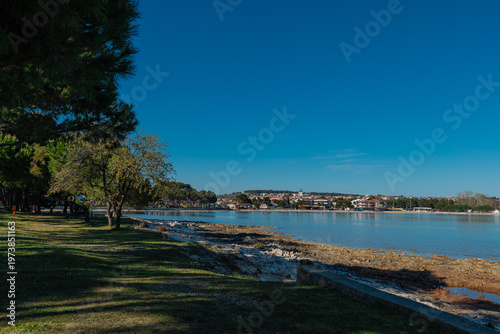 Coastal landscape in Medulin with calm sea, rocky shoreline and green park under clear blue sky, capturing peaceful seaside atmosphere, natural beauty and relaxed Mediterranean lifestyle.